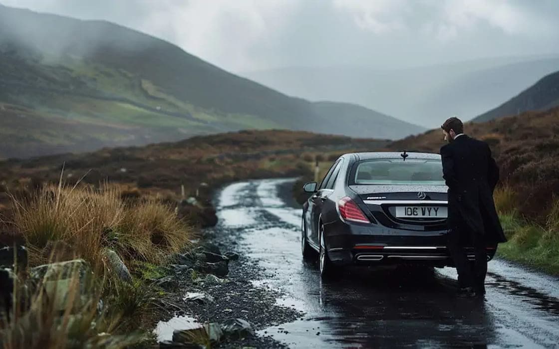 A chauffeur beside an executive saloon on a mountain road in the rain
